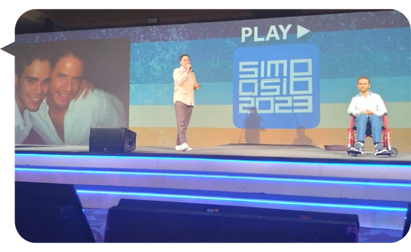 Kike y Gino en el escenario durante el Simposio 2023, con una imagen proyectada de ellos y el logo del evento.