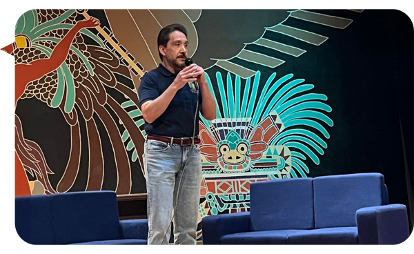 Ness Carballo, conferencista internacional, hablando en un escenario decorado con arte de estilo mesoamericano, mientras sostiene un micrófono.