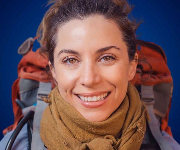 Fernanda Pérez Alarcón, mujer sonriente con mochila y bufanda, vestida para una expedición al aire libre, con un fondo azul.