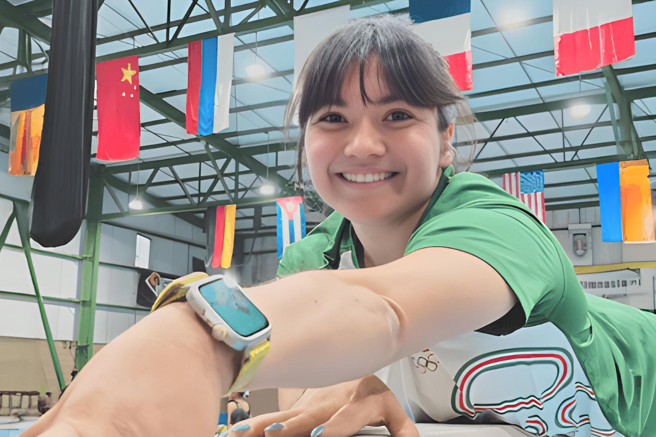 Alexa Moreno sonríe mientras entrena en un gimnasio internacional con banderas al fondo, promoviendo el deporte juvenil en la Believe Cup 2025.