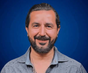 Fotografía de Daniel Villarreal, sonriendo con una camisa gris clara sobre un fondo azul, proyectando seguridad y cercanía.